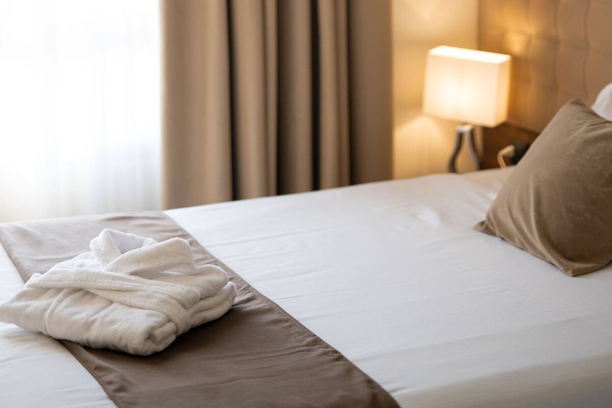 Ein Hotelzimmer mit einem ordentlich bezogenem Bett, einer Lampe auf dem Nachttisch und einem Fenster mit Vorhängen.