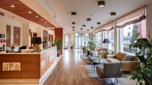 A spacious lobby with a wooden reception desk, comfortable seating area, large windows, and indoor plants. - Charme (Prato)
