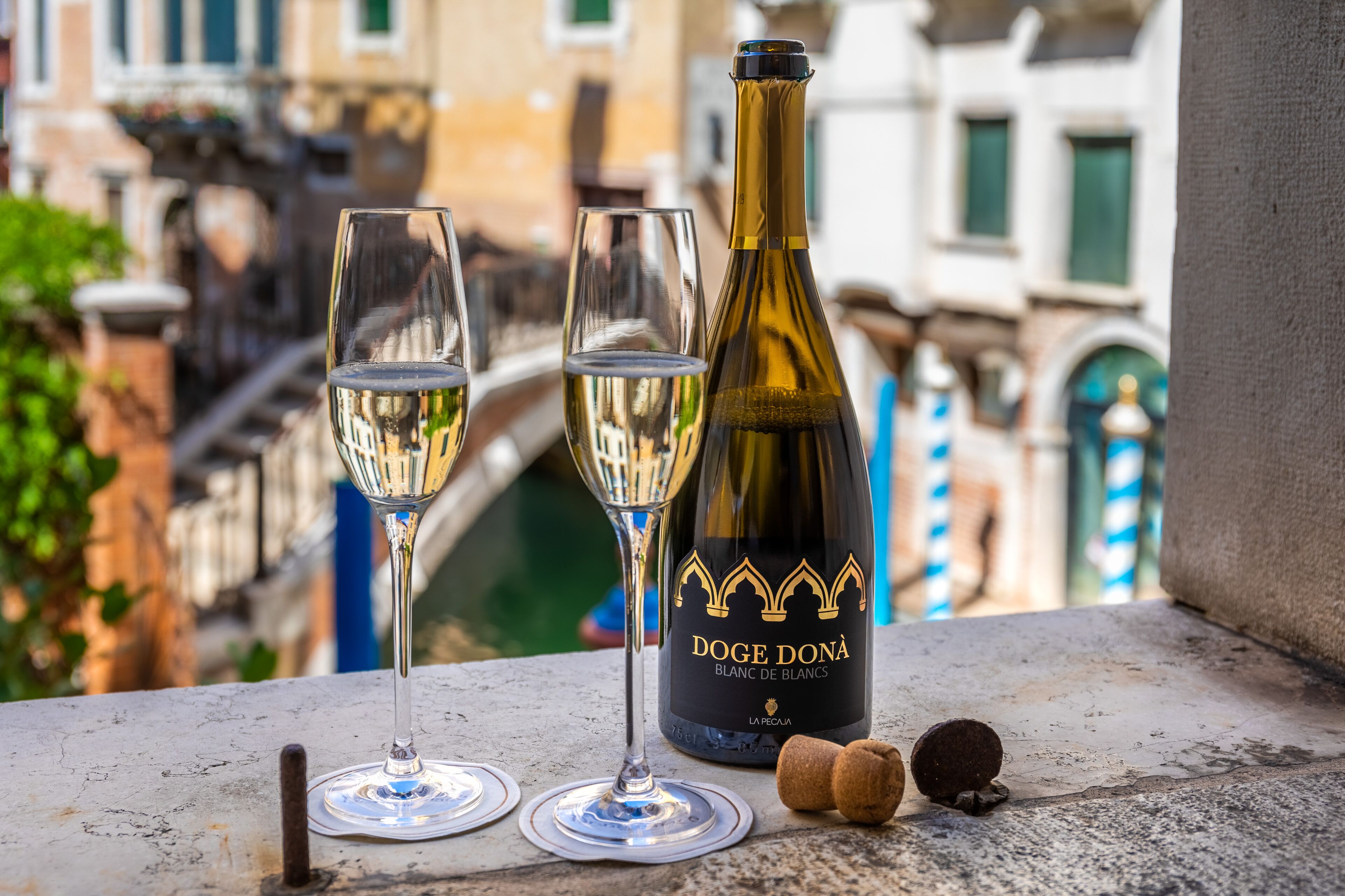 Una bottiglia di vino e due bicchieri su un balcone con vista su un canale e palazzi.