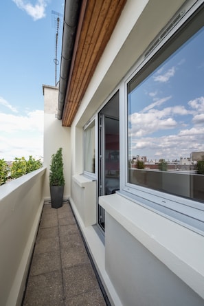 A narrow balcony with a potted plant, a window with a city view, and a wooden ceiling.