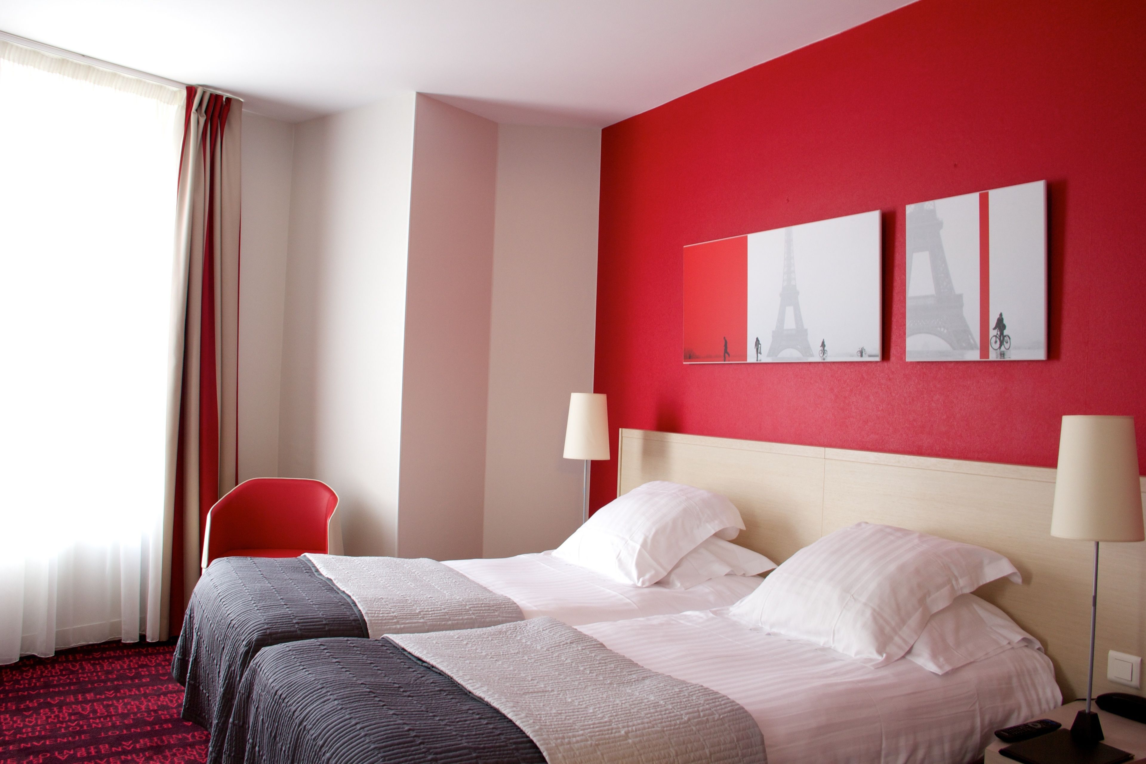 Quarto de hotel com uma parede de destaque vermelha, duas camas, uma cadeira vermelha e uma obra de arte da Torre Eiffel.