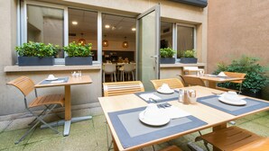 An outdoor dining area with tables and chairs, potted plants, and a view of the interior through a glass door. - Hôtel Le Richemont (Paris)