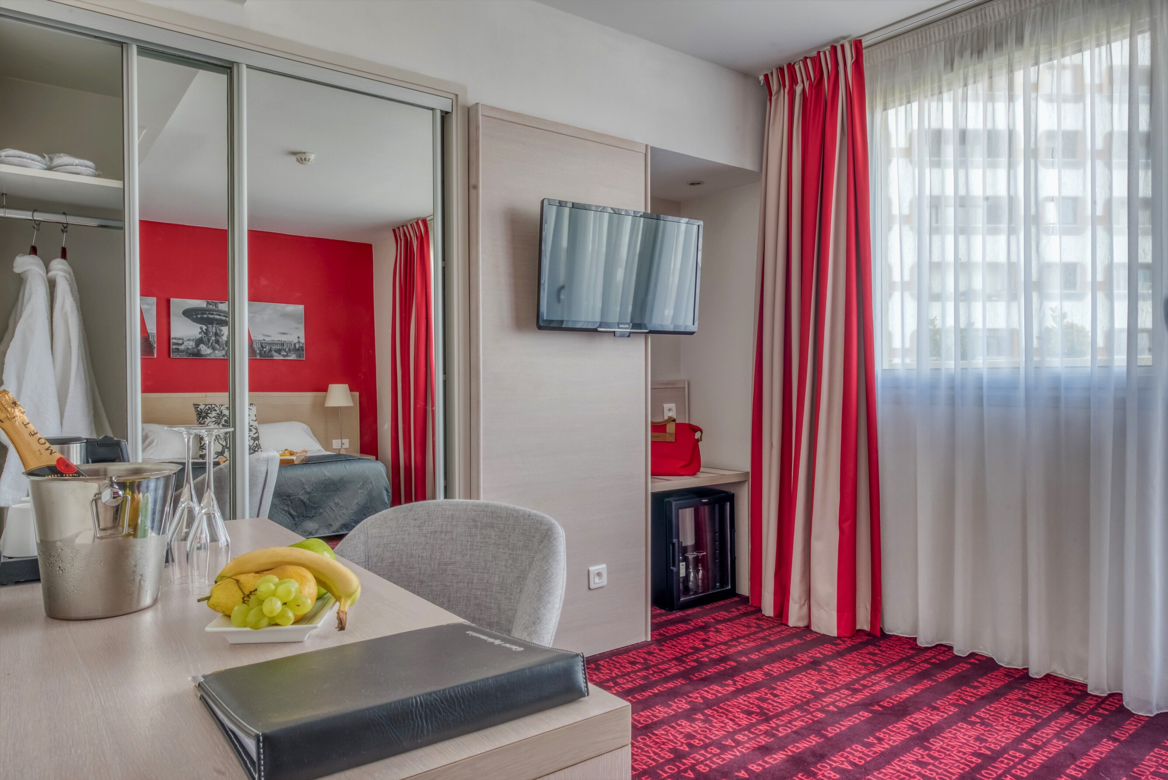 A hotel room with a bed, a desk with a laptop, a TV, a minibar, and a red and white striped curtain.