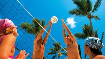 Un partido de voleibol de playa con jugadores que se estiran para alcanzar la pelota, palmeras y un cielo azul despejado.