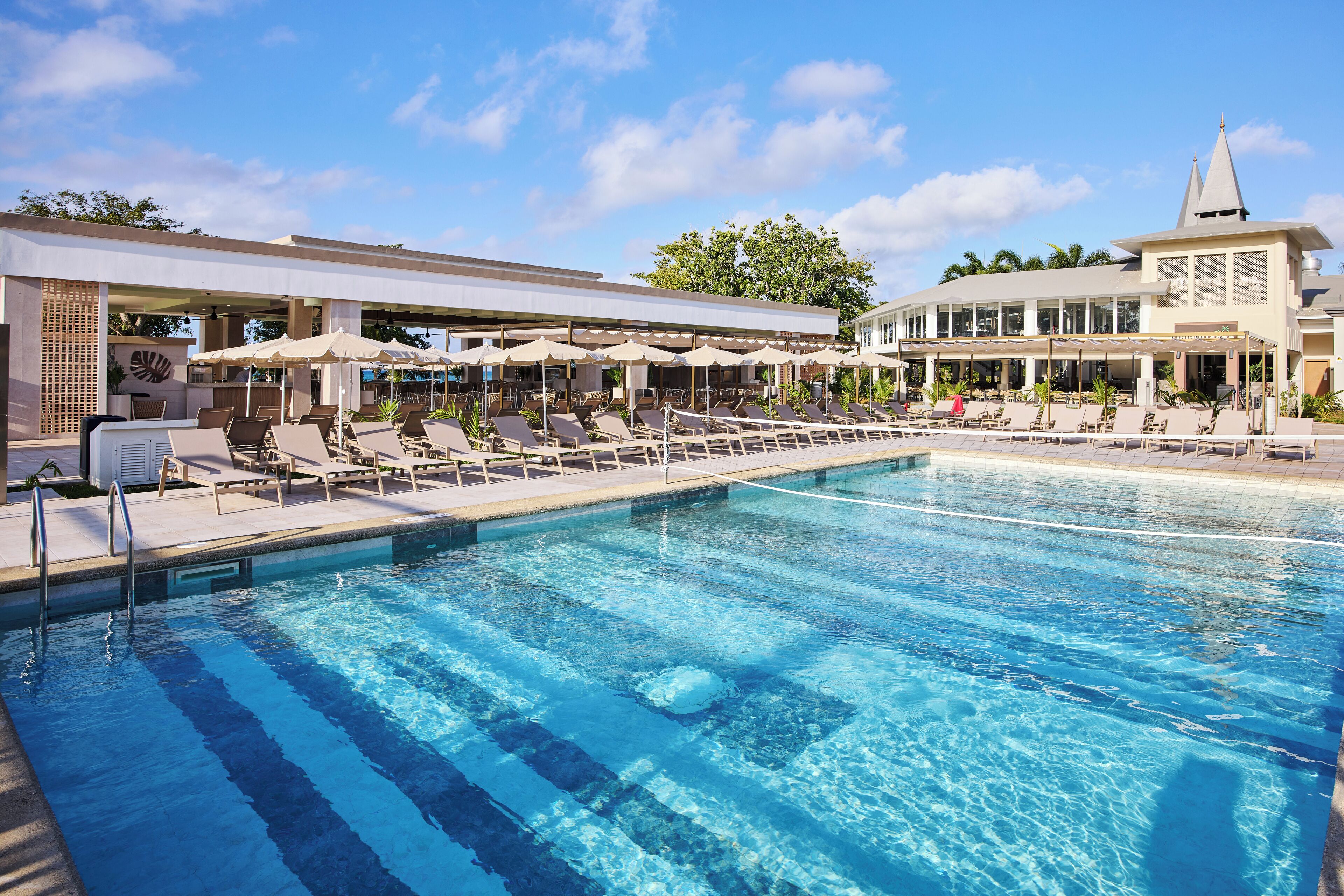 Une piscine entourée de chaises longues et de parasols, le tout bordé d’un bâtiment à l’architecture moderne et d’une tour.