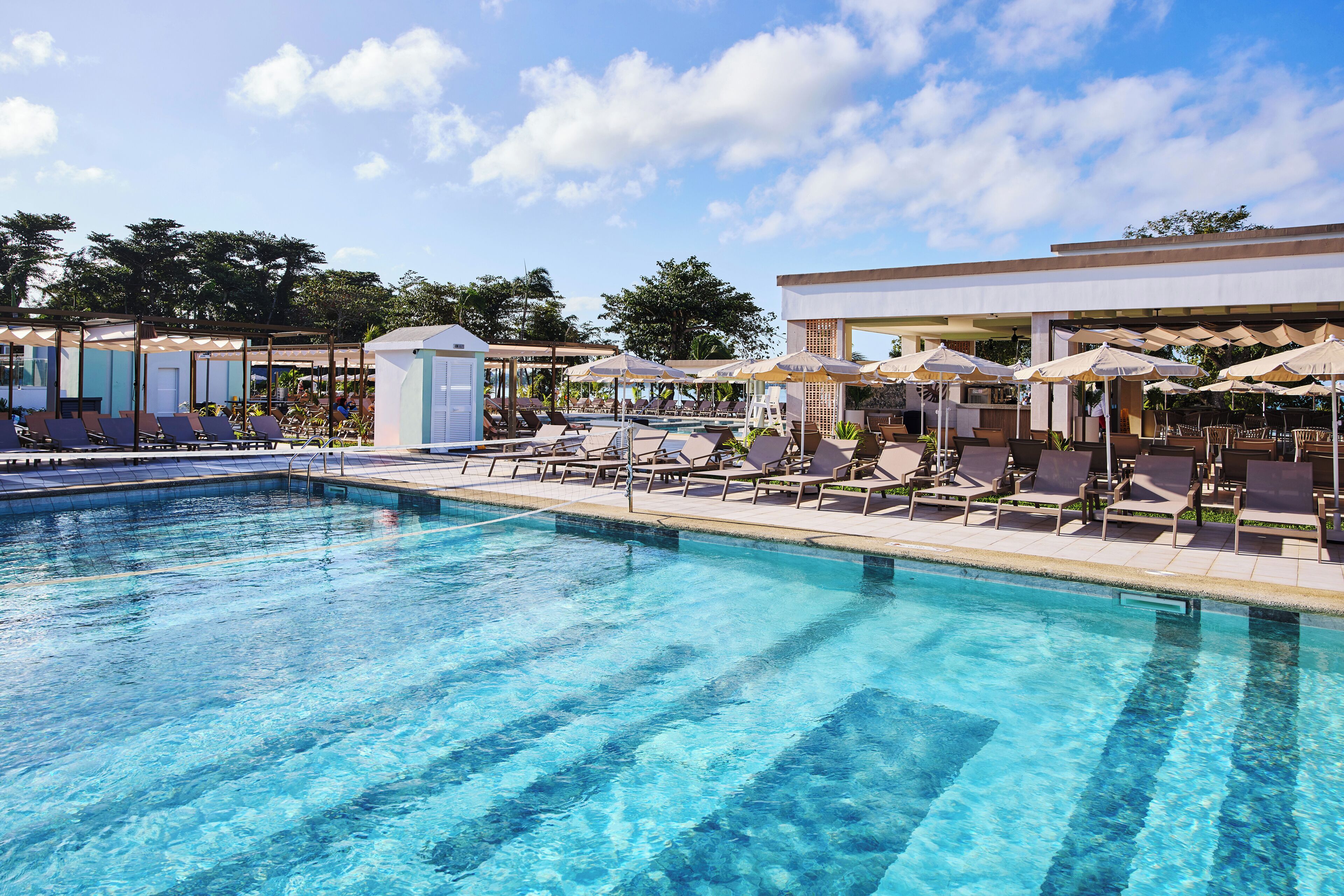 Un área de piscina con sillas de descanso y sombrillas, rodeada de edificios y árboles.