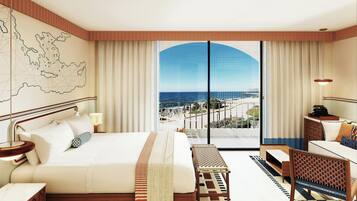 A hotel room with a large bed, a desk, a chair, a map on the wall, and a view of the sea through a large window.