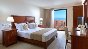 Una camera d'albergo con un letto grande, due comodini con lampade, una scrivania in legno con un televisore e un balcone vista mare.