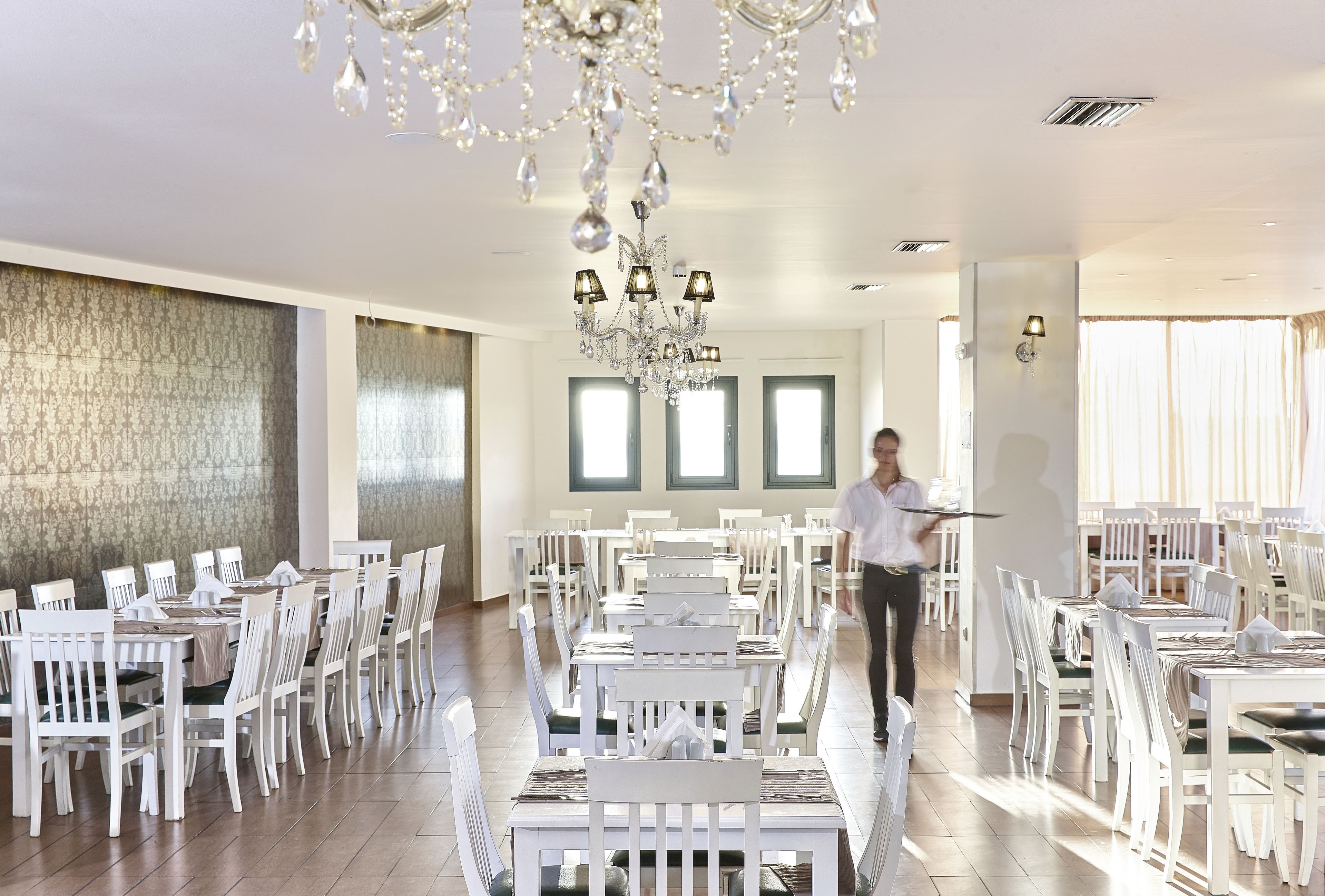 A spacious dining area with white tables and chairs, a chandelier, and large windows.