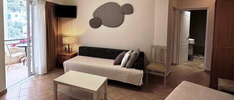 A hotel room with a bed, a sofa, a table, a TV mounted on the wall, and a balcony with a chair.