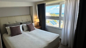 A hotel room with a large bed, a nightstand with a lamp, and a view of the ocean through a window. - Faedra Beach Hotel (Agios Nikolaos)