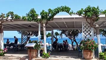 A beachside café with outdoor seating, a pergola structure, and a view of the sea.