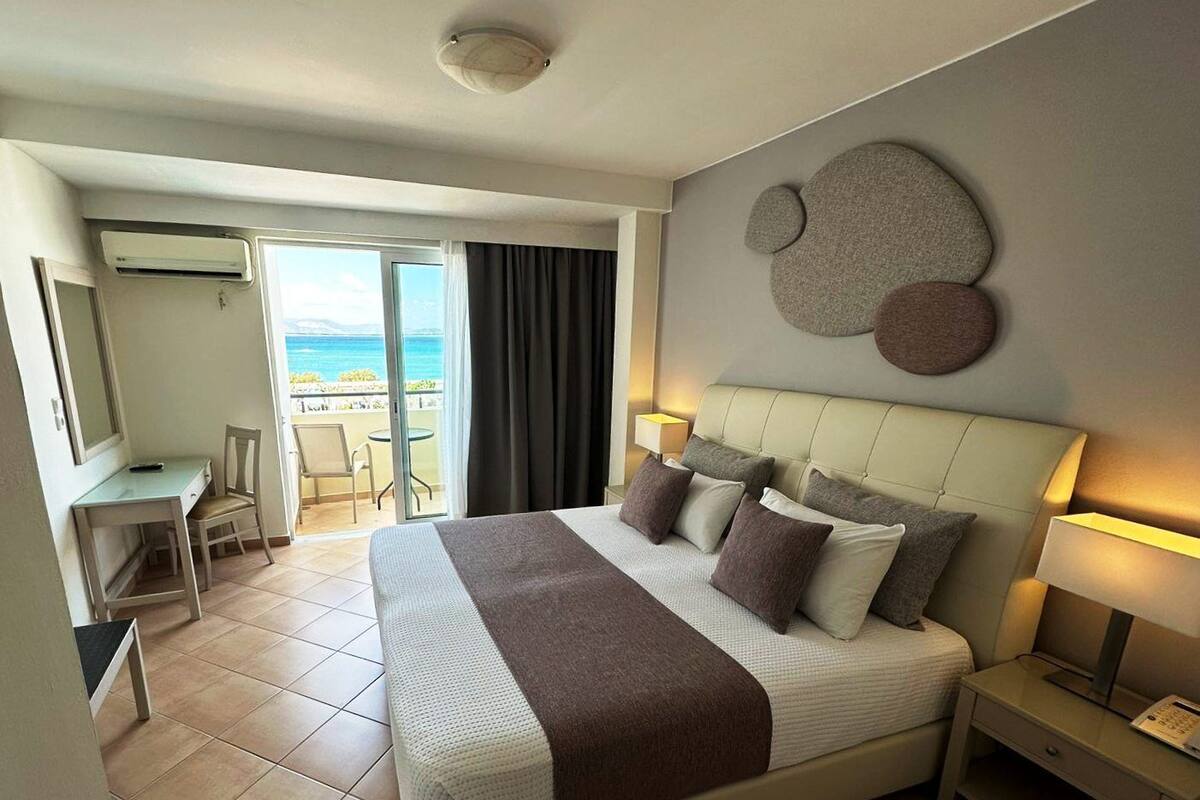 Habitación de hotel con cama, mesita de noche, escritorio, silla y vista al mar a través de una puerta corrediza.