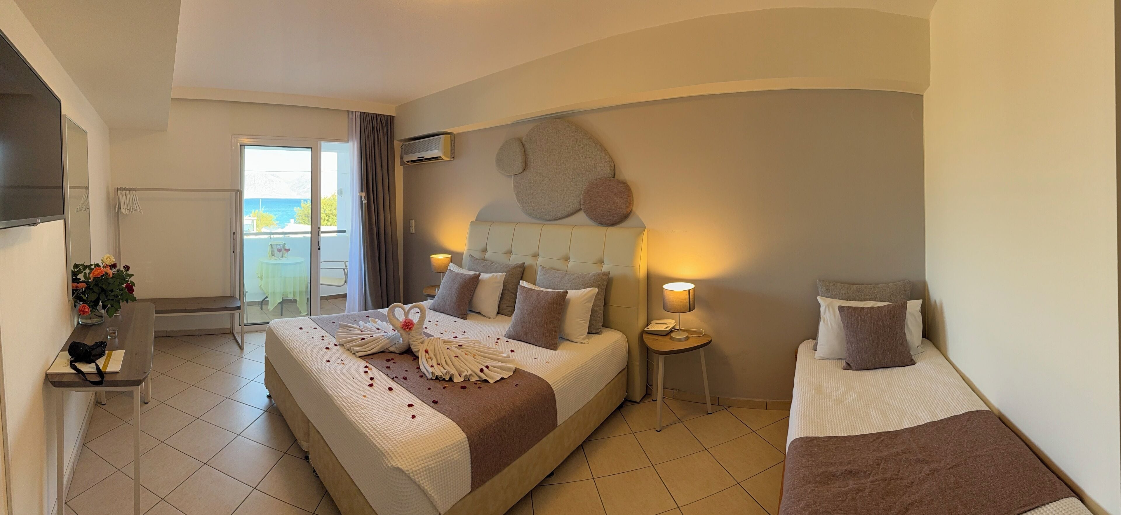 Une chambre d’hôtel avec un grand lit, un lit plus petit, un bureau avec une lampe et une vue sur la mer, accessible par une porte coulissante.