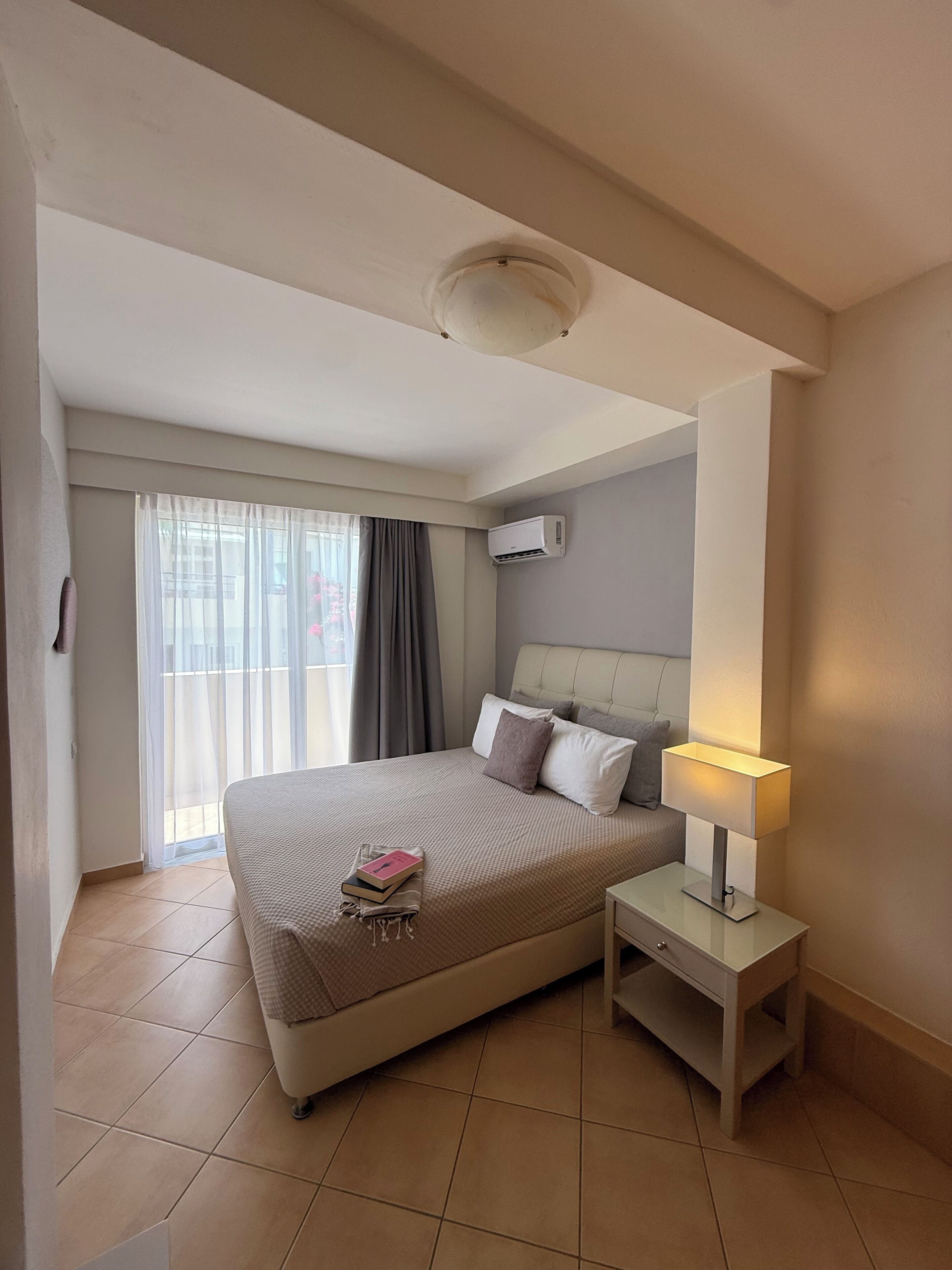 A hotel room with a bed, a bedside table with a lamp, an air conditioning unit, and a window with curtains.