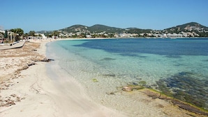 A beach with clear water, a sandy shore, and buildings in the background.