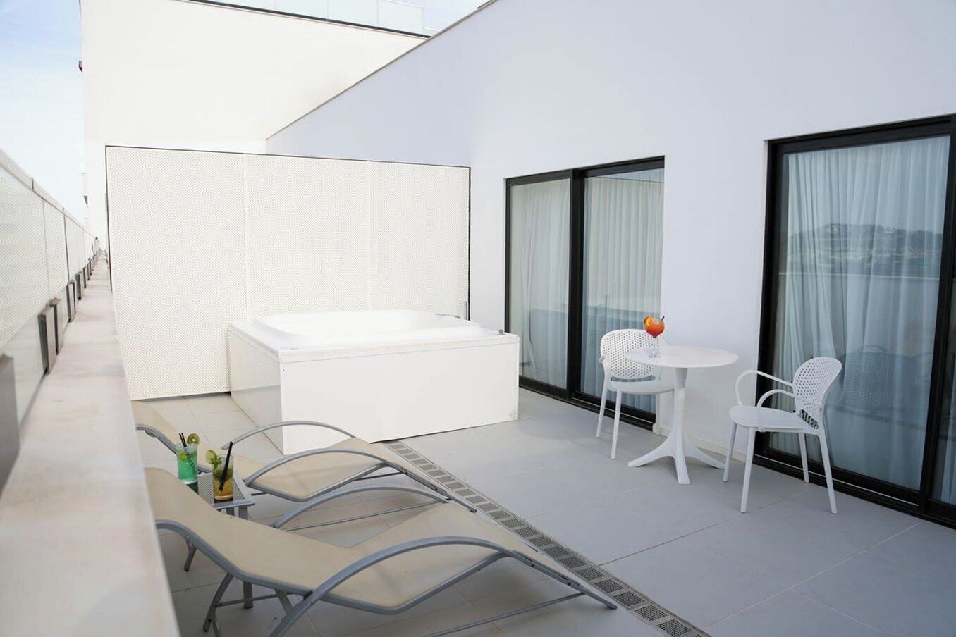 A modern outdoor balcony with a white sofa, two white chairs, a small round table, and a glass partition.