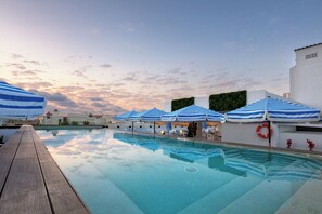 A swimming pool with blue and white striped umbrellas, a wooden deck, and a white building with a green hedge. - Eurostars Puerto de Ibiza (Ibiza Town)