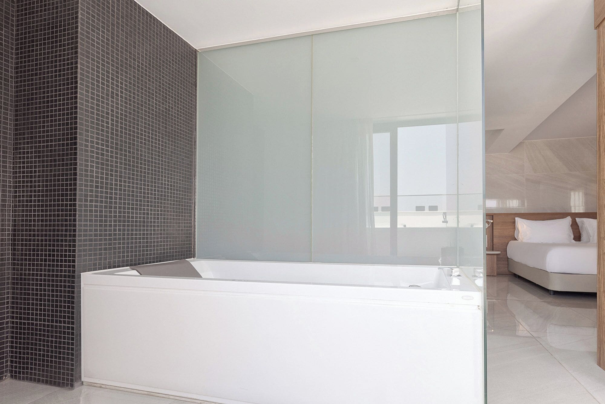 A modern bathroom with a large white bathtub, a glass shower enclosure, and a view of a bedroom with a bed and a nightstand.
