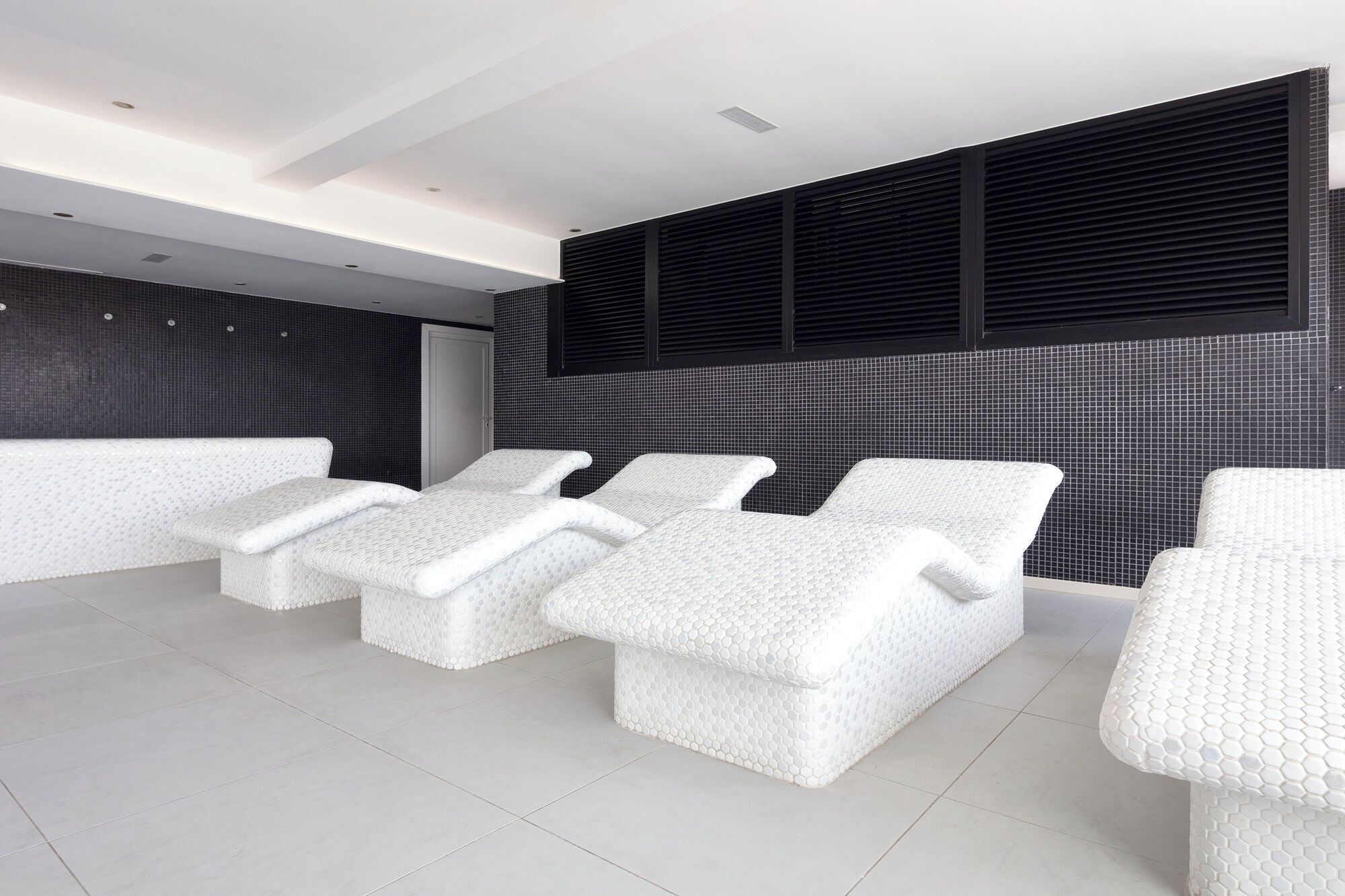 Un spa moderno con tres sillones blancos y texturizados, y un ventanal grande con persianas negras.