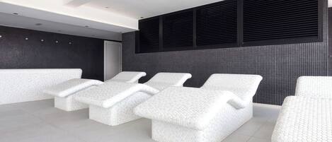 Un spa moderno con tres sillones blancos y texturizados, y un ventanal grande con persianas negras.