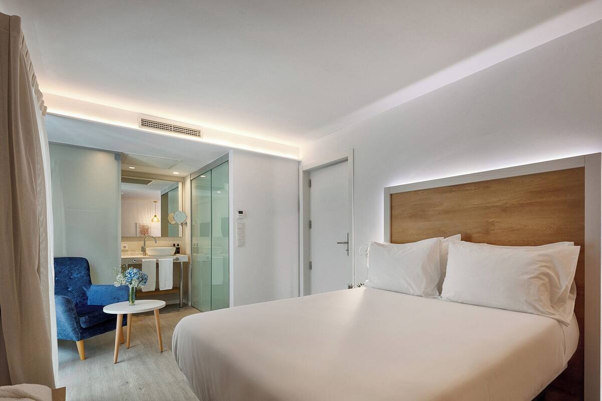 Une chambre d’hôtel avec un grand lit, une petite table ronde, une chaise bleue et une salle de bain visible à travers une porte ouverte.