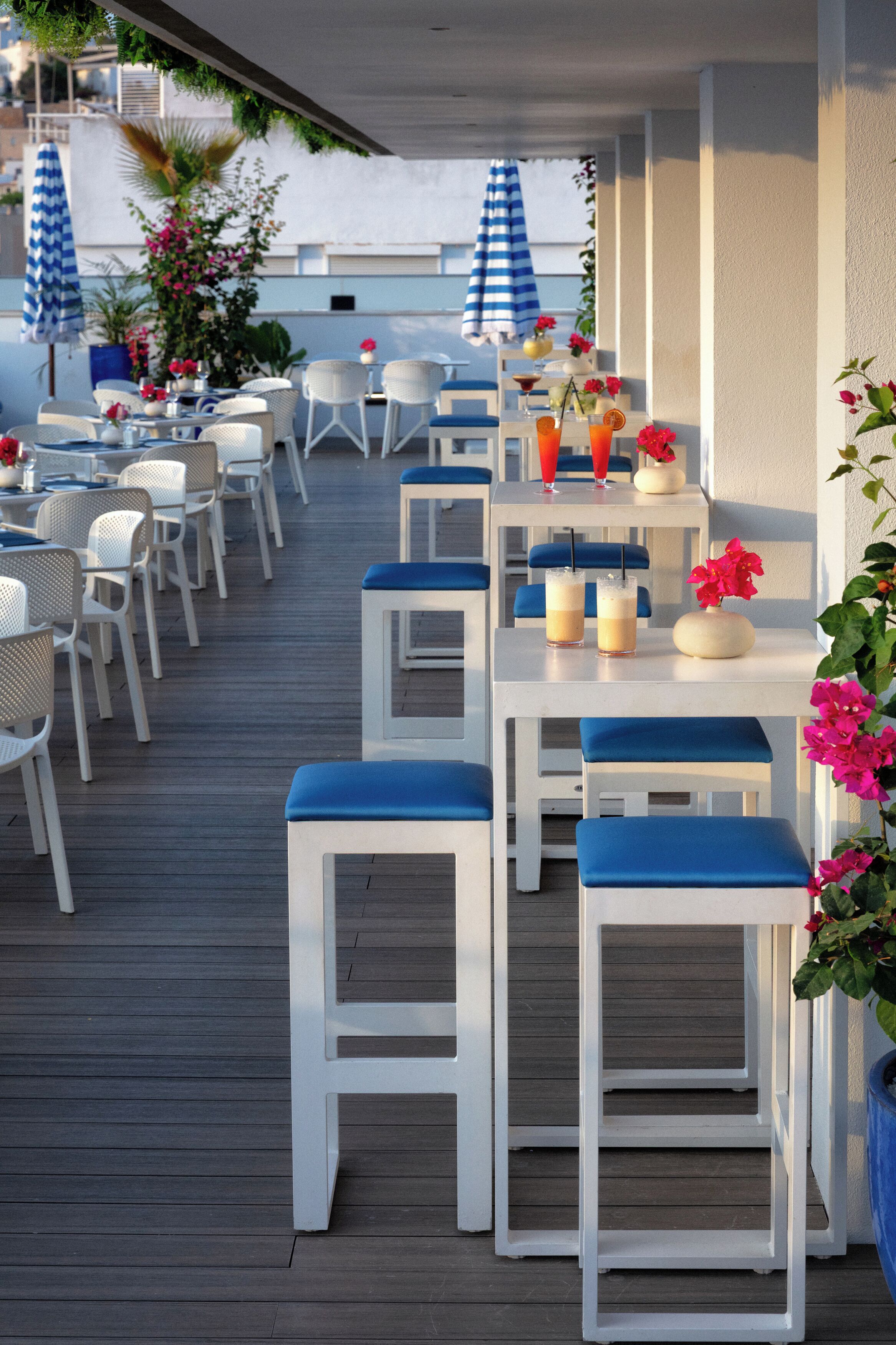 Eine Terrasse im Freien mit weißen Tischen und blauen, gepolsterten Hockern, die auf einer Holzterrasse angeordnet sind.