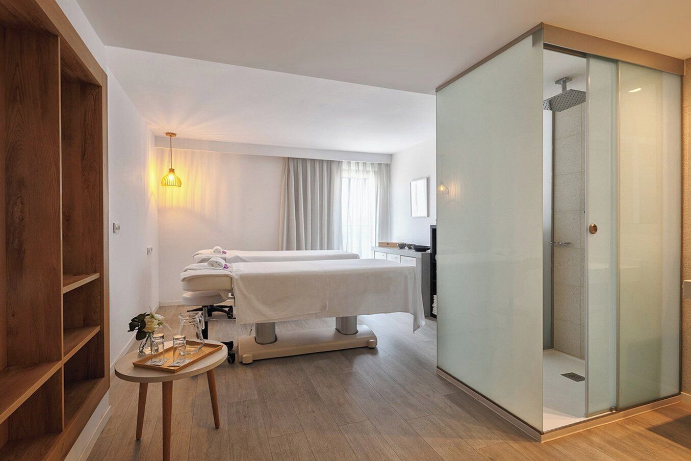 A hotel room with a massage table, a small table with a vase, a shower area, and a wooden shelf.