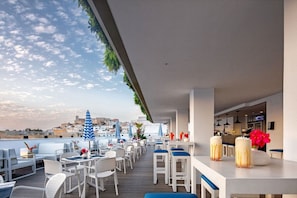 An outdoor seating area with white tables and chairs, blue-striped umbrellas, and a view of the cityscape in the background.