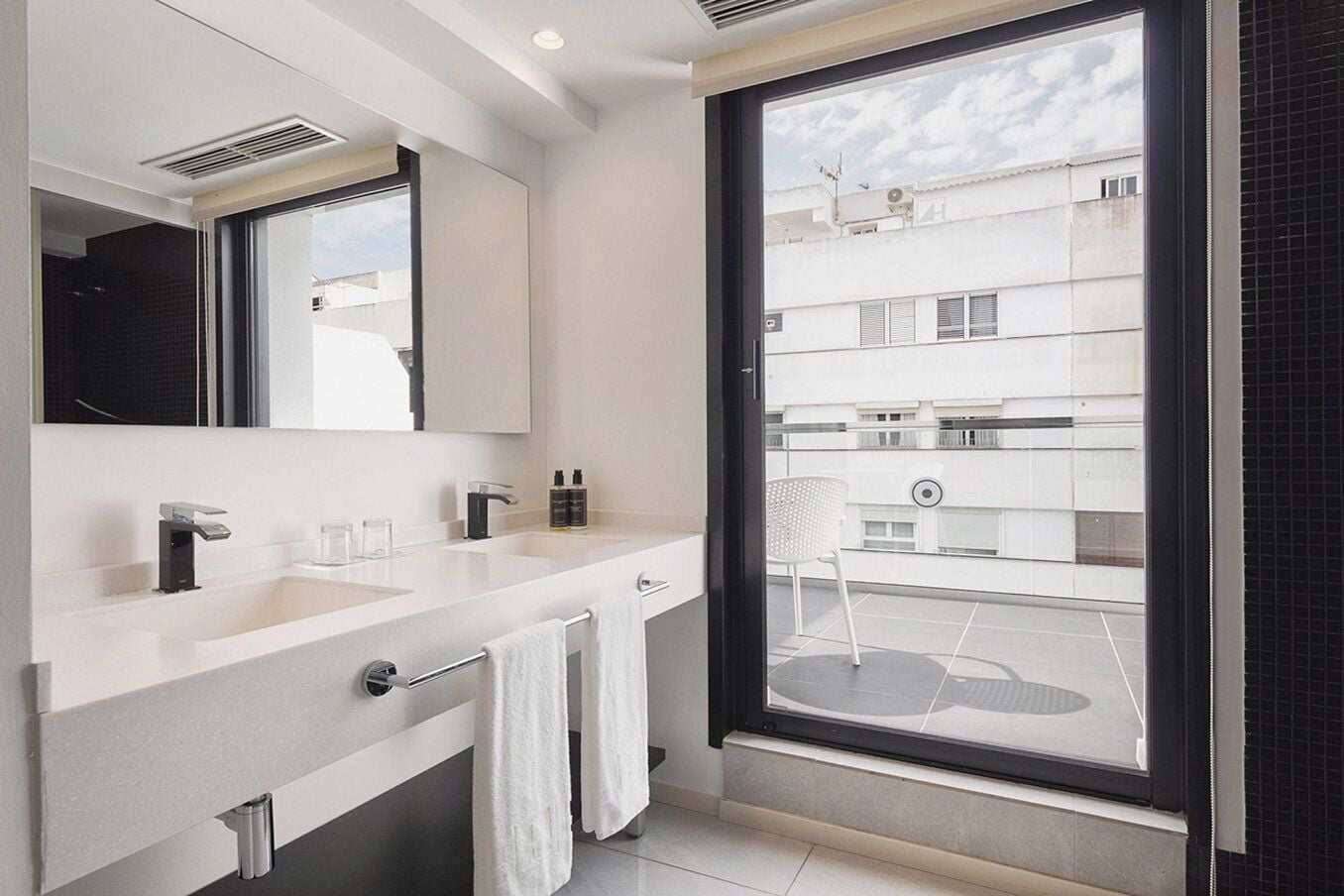 A modern bathroom with a double sink, a mirror, and a view of a balcony with a chair.
