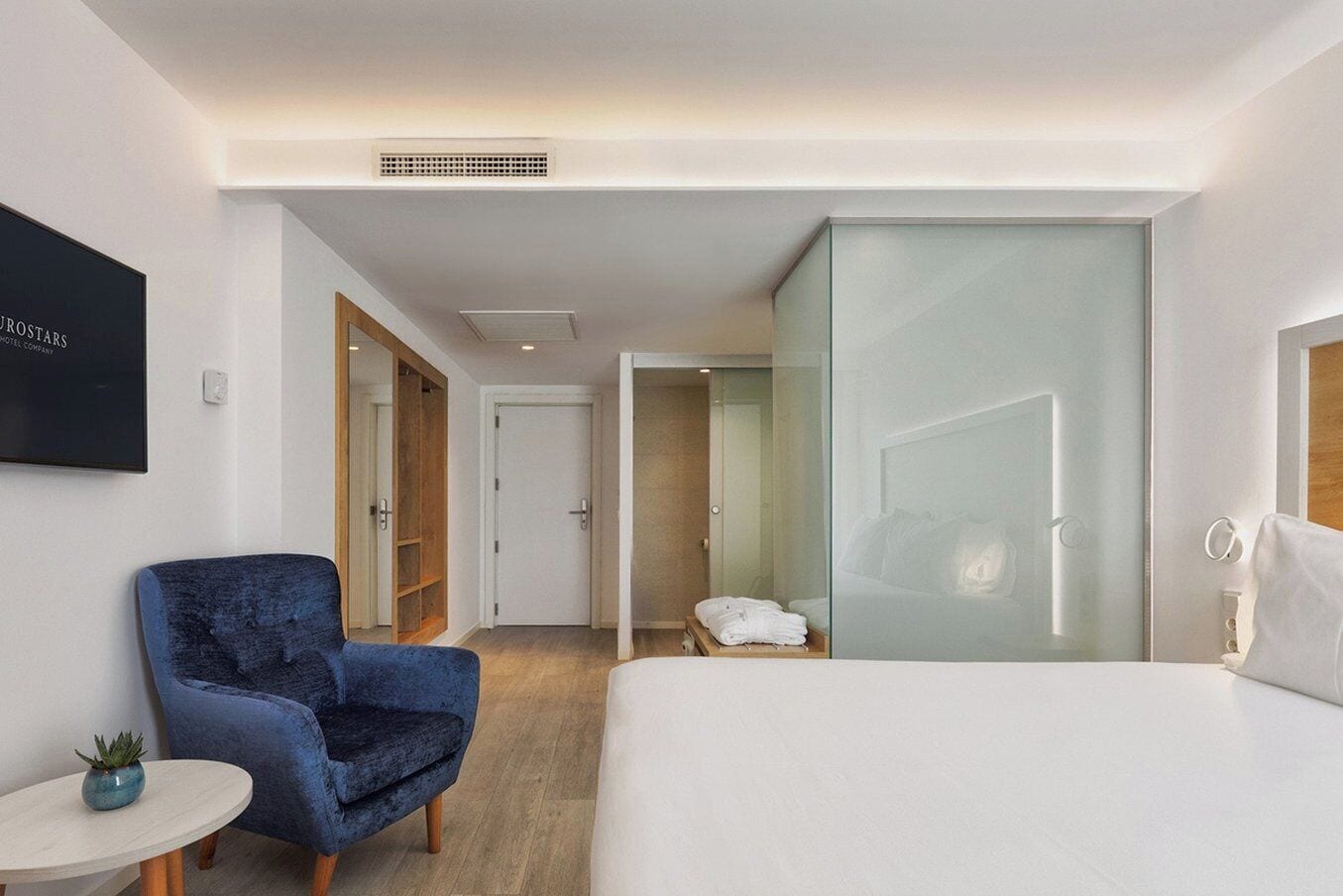 A hotel room with a bed, a blue armchair, a small table with a plant, a glass partition, and a door leading to another room.
