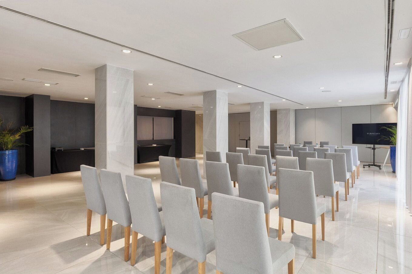 Un salón de conferencias con filas de sillas grises, una pantalla grande y una recepción al fondo.