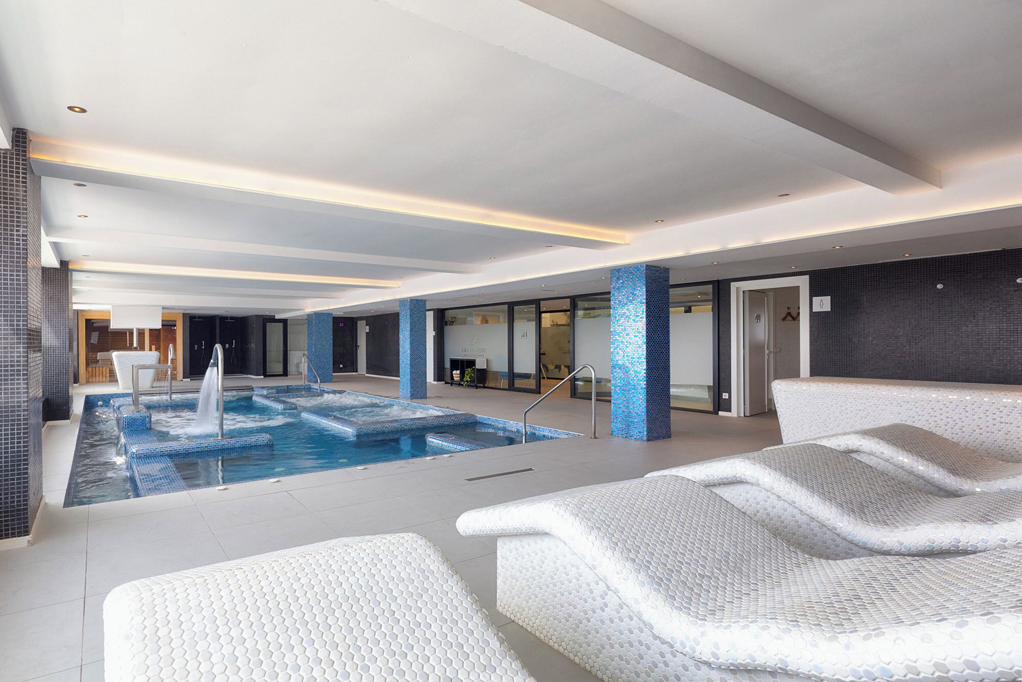 A modern indoor pool area with a central hot tub, seating areas, and a reception desk.