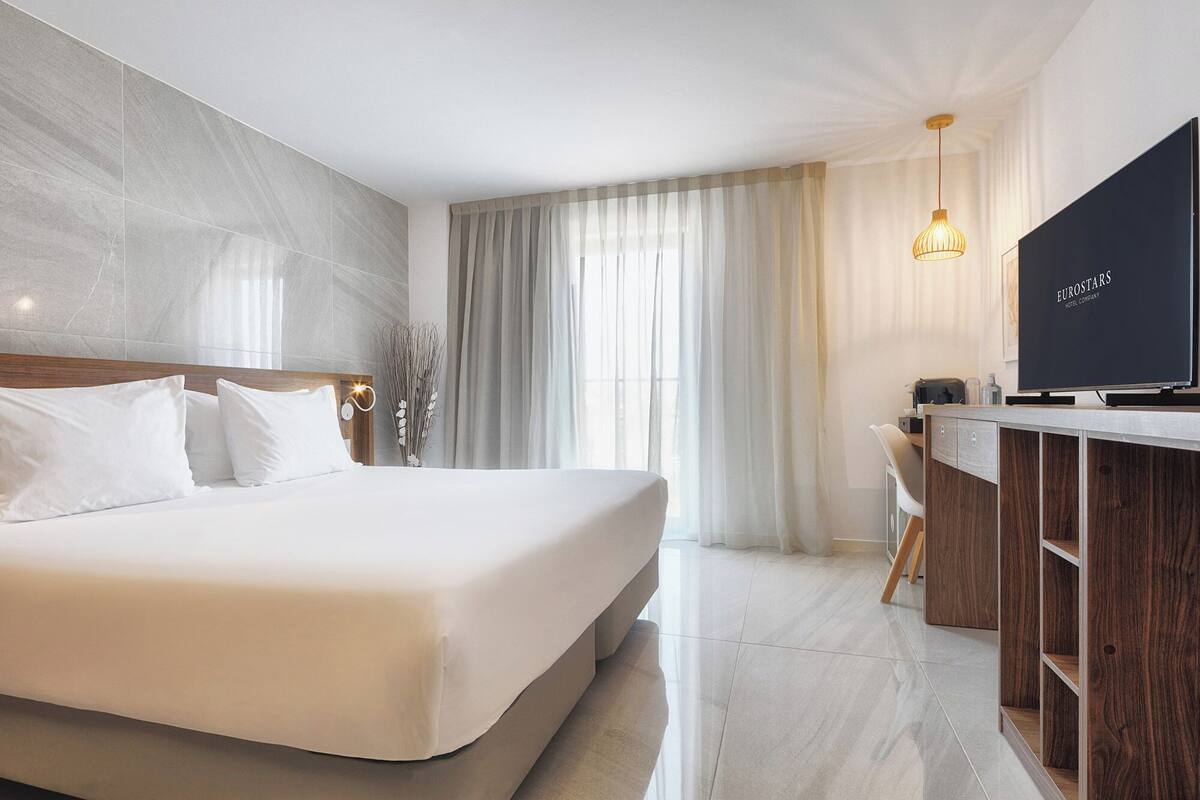 Une chambre d’hôtel moderne dotée d’un grand lit, d’un bureau avec un téléviseur à écran plat et offrant une vue sur la ville à travers de fines rideaux.