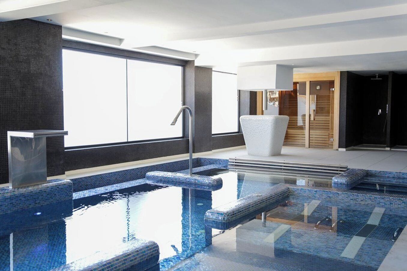 A modern indoor pool area with clear glass walls, a metal pool ladder, and a white lounge chair.