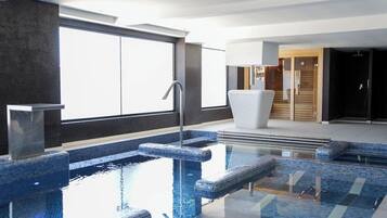 A modern indoor pool area with clear glass walls, a metal pool ladder, and a white lounge chair.