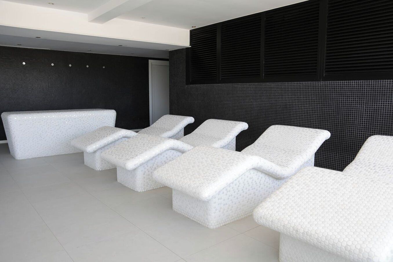 A room with a row of white, textured lounge chairs aligned against a dark wall with a grid pattern.