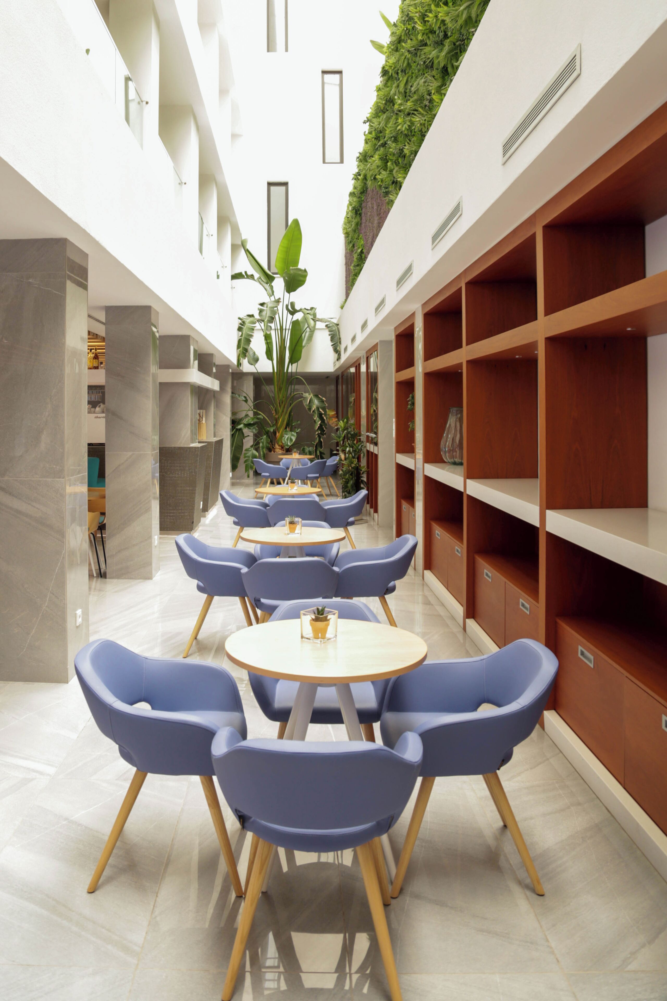 Een moderne binnenruimte met een centraal zithoekgedeelte met ronde tafels en blauwe stoelen, omringd door houten planken en een groene verticale tuin.