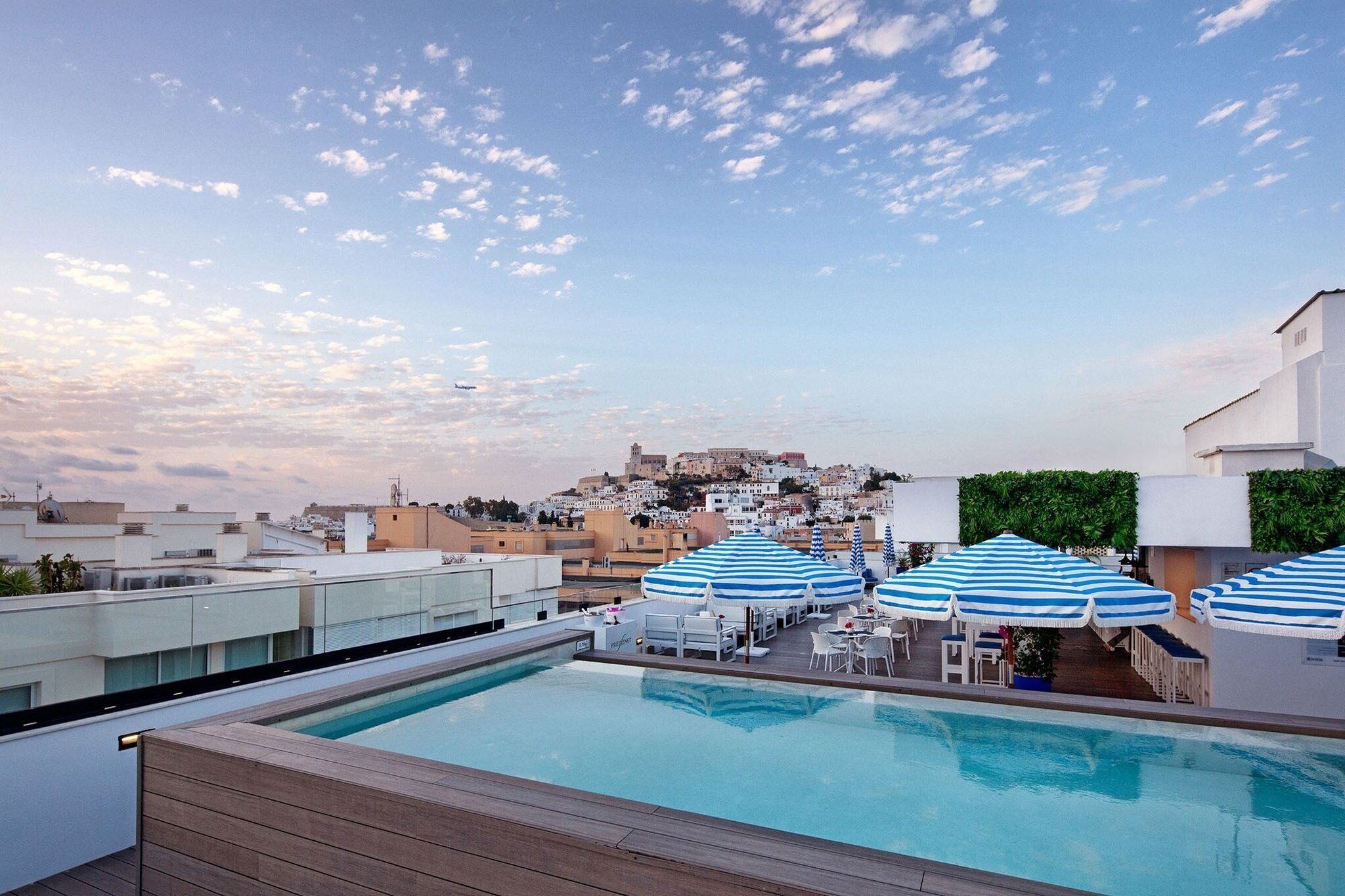Una piscina en la azotea con sombrillas azules a rayas, vista de un paisaje urbano y cielo despejado.