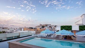 Una piscina en la azotea con sombrillas azules a rayas, vista de un paisaje urbano y cielo despejado.