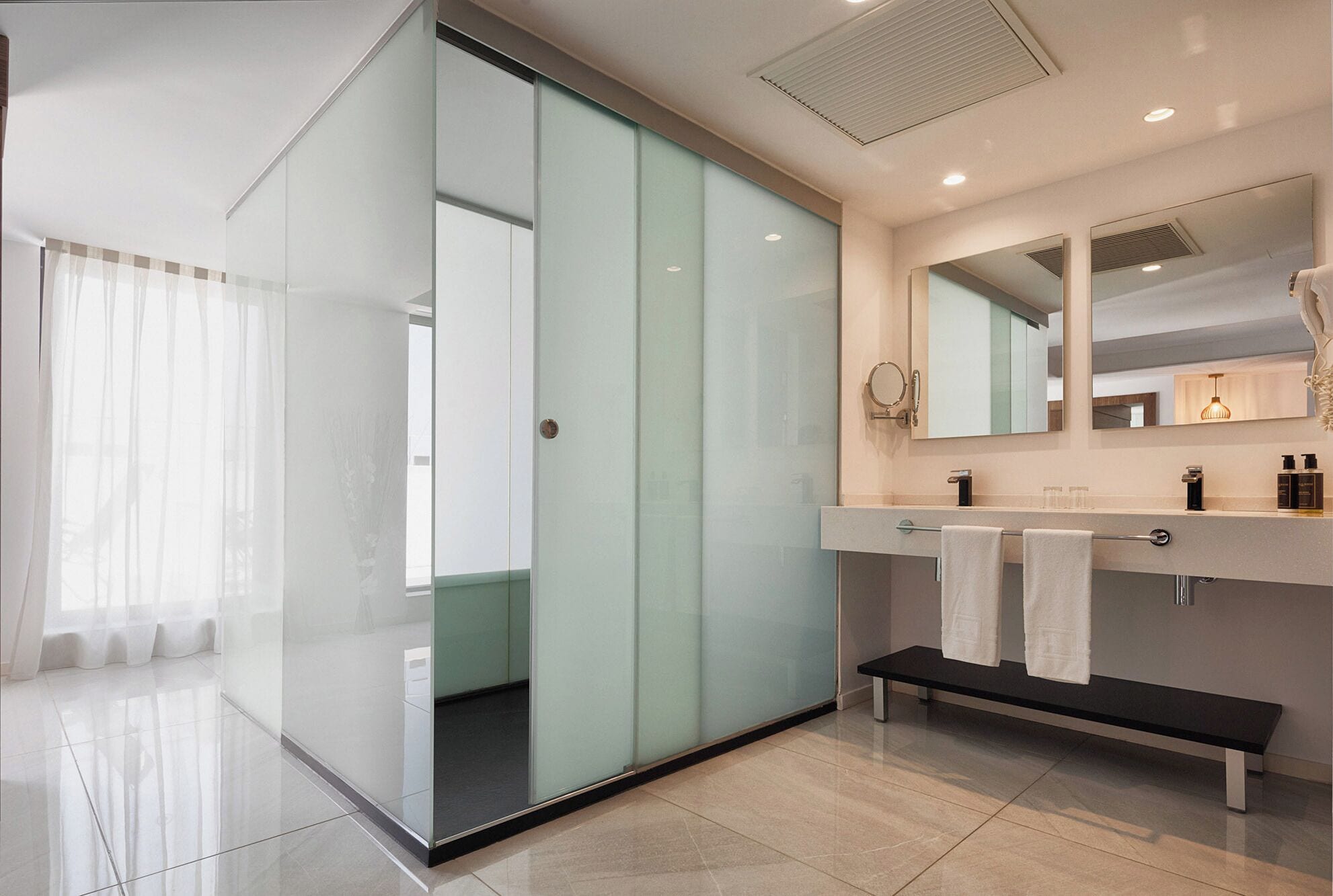 A modern bathroom with a glass partition, a bench, and a vanity with a mirror and towel rack.