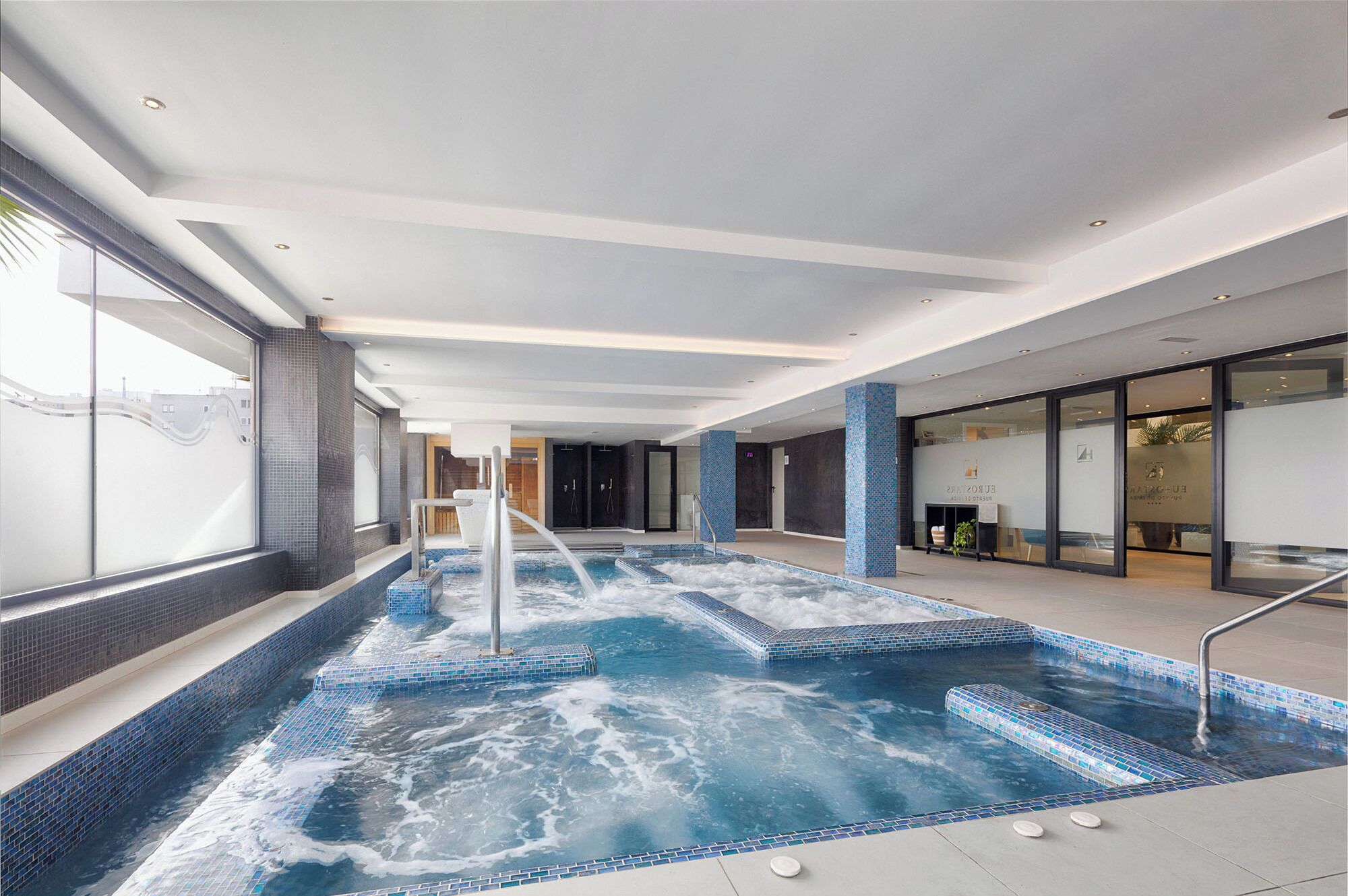 A modern indoor pool with clear water, a central jet, and a glass wall on one side.