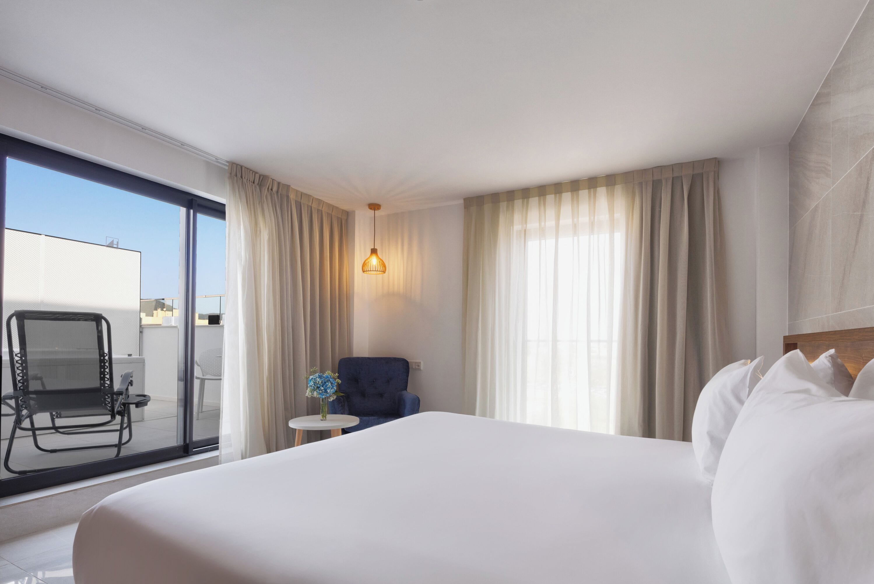 A hotel room with a large bed, a chair, a small table with a vase, and a view of the outside.