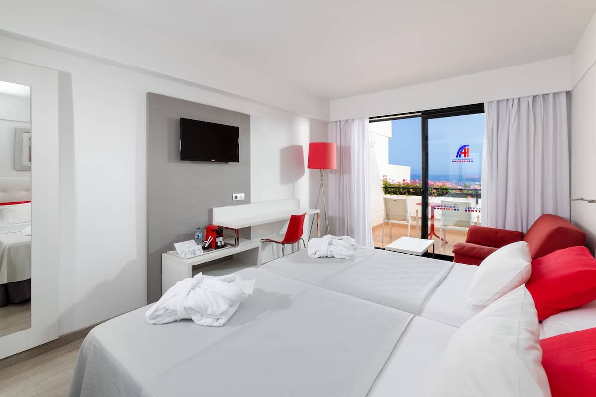 A hotel room with a large bed, a flat-screen TV mounted on the wall, a red chair, a small table with a lamp, and a view of the outdoors through a large window.