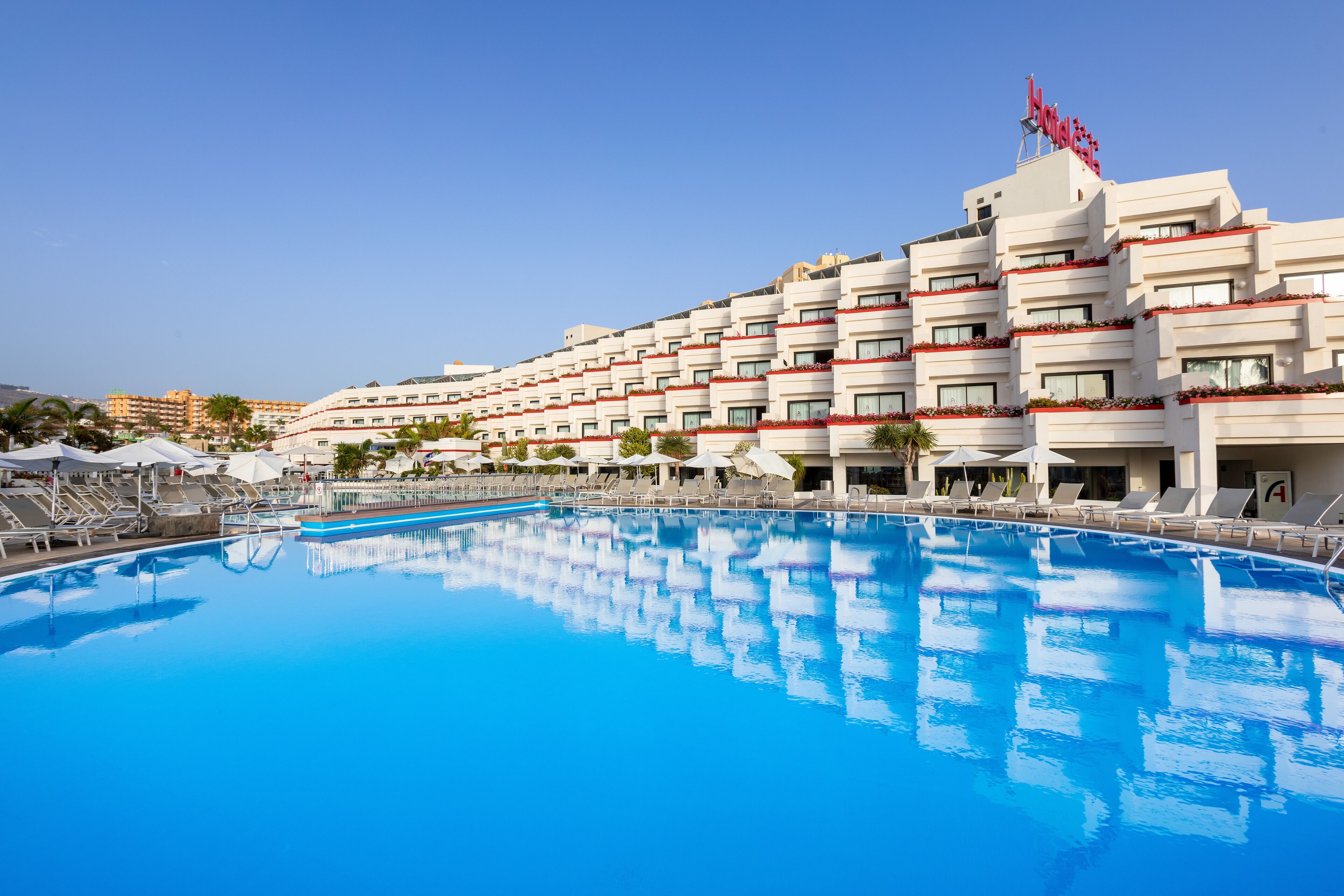 Un hotel con una amplia piscina, varios balcones y un cielo azul despejado.