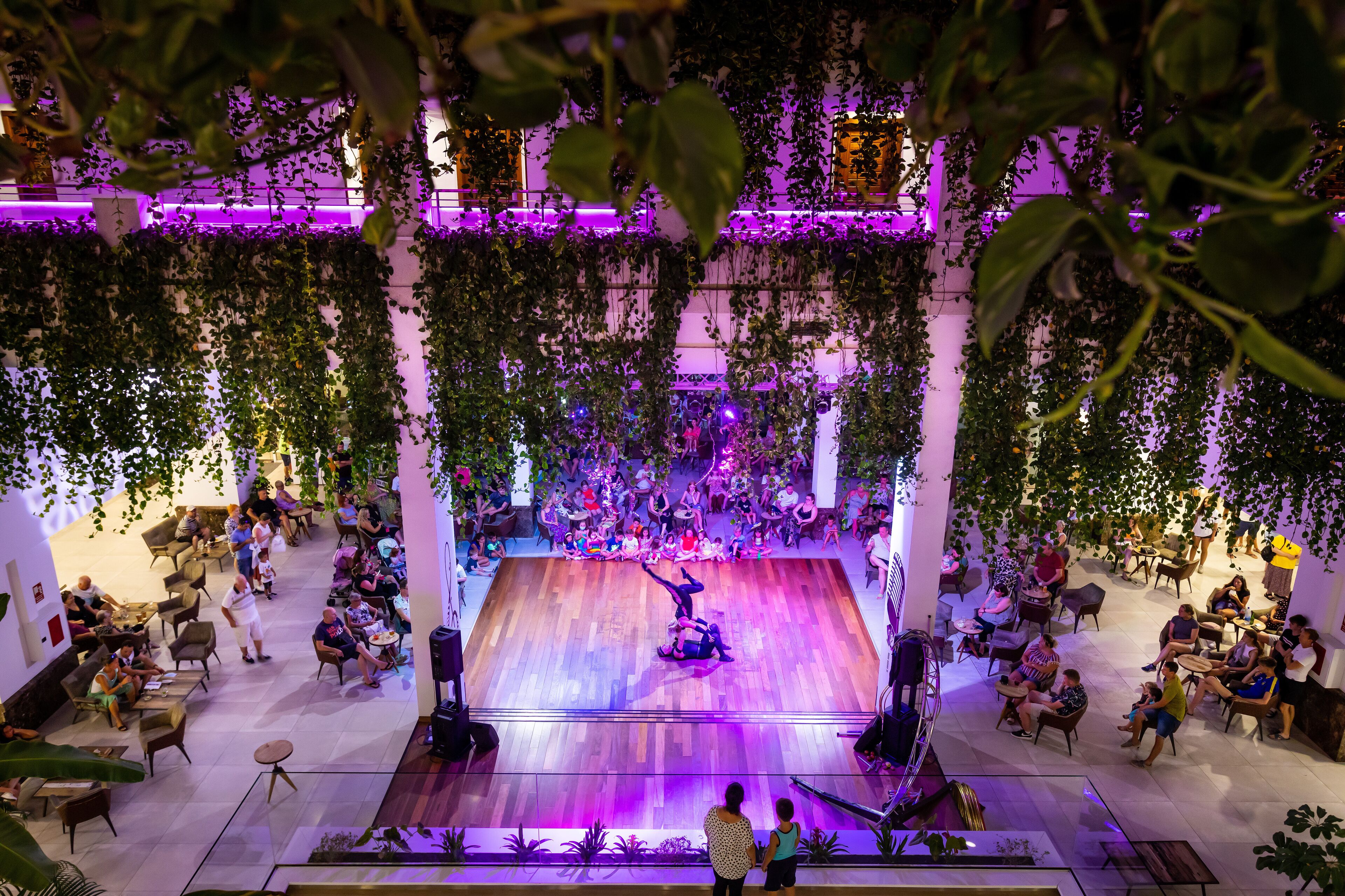 A spacious indoor venue with a central stage, surrounded by seating areas and lush greenery.