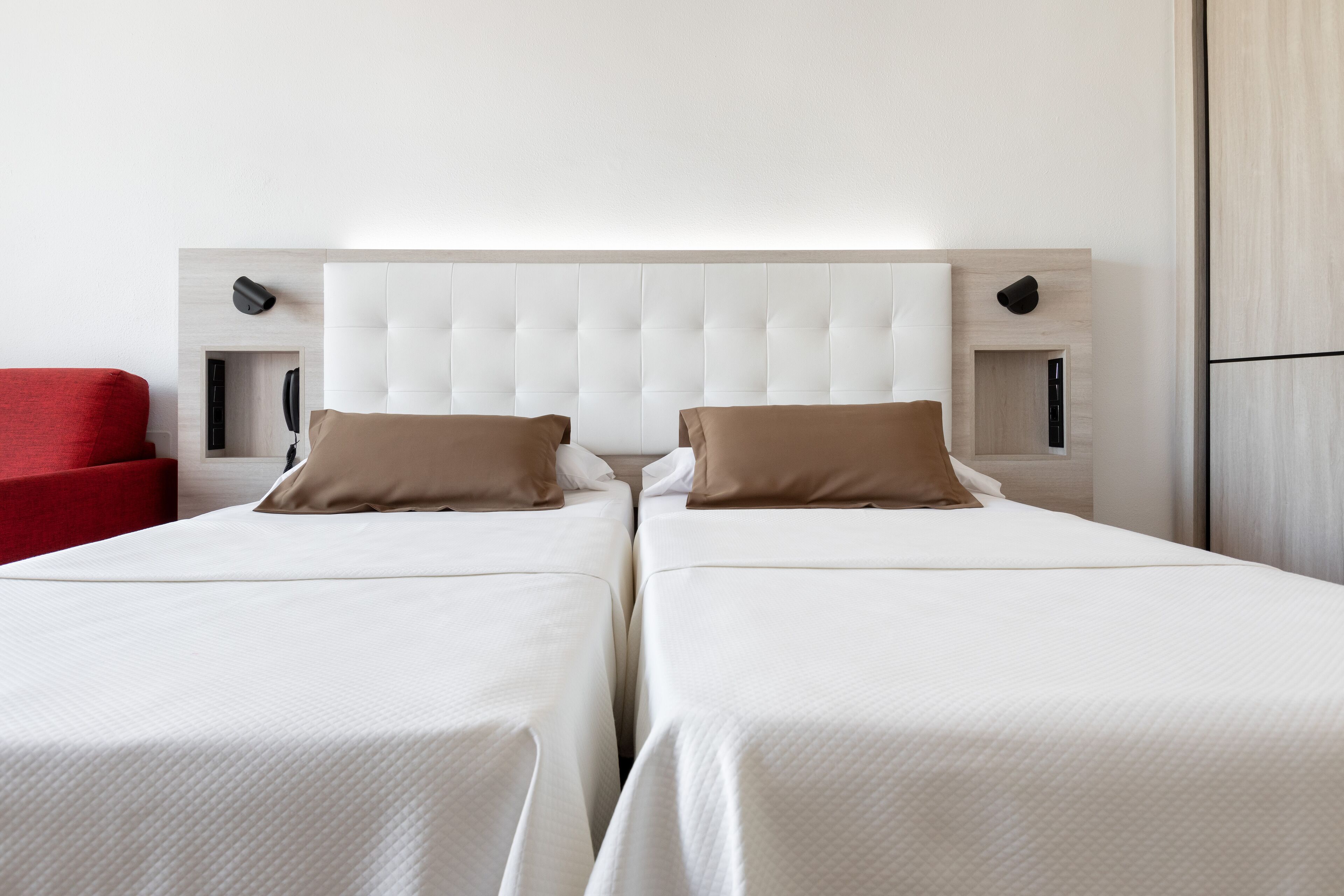 Habitación de hotel con dos camas, una silla roja y un cabecero moderno.