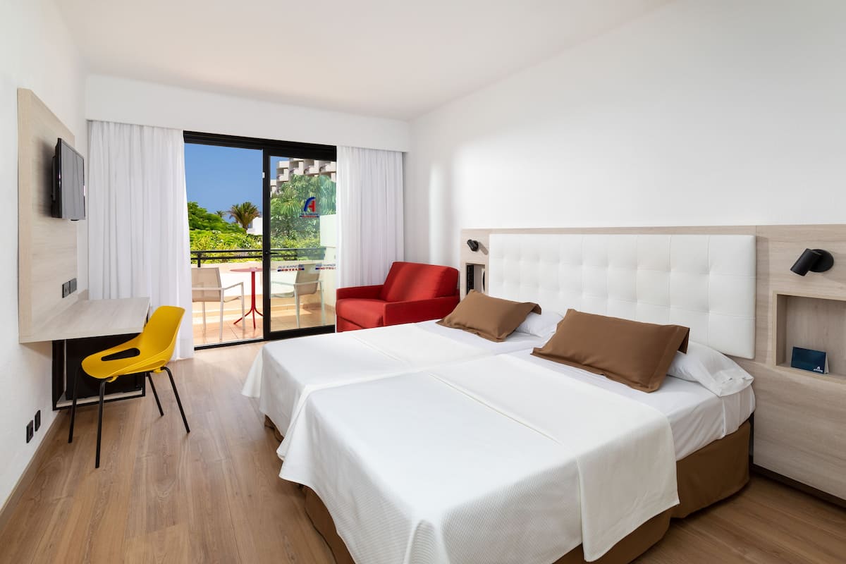 A hotel room with a large bed, a red chair, a TV, and a balcony with a view of greenery.