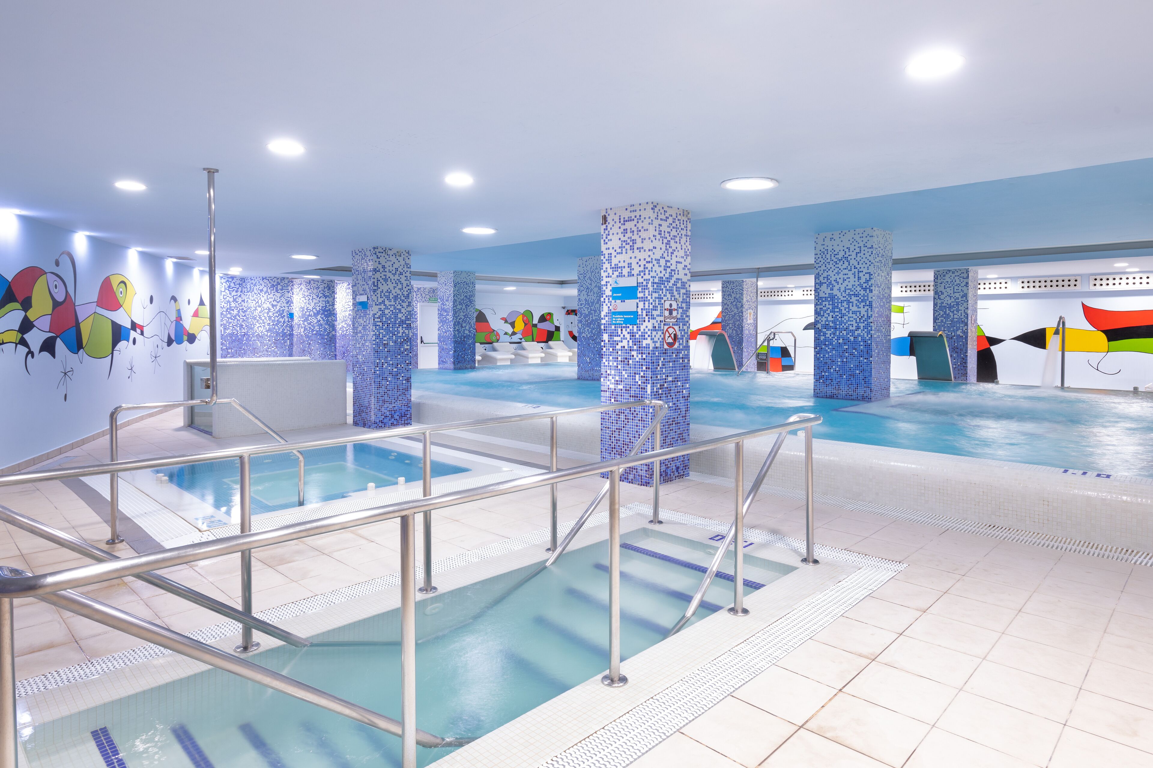Una piscina interior con paredes de azulejos, barandillas de metal y coloridos adornos murales.