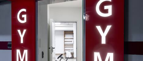 A gym entrance with illuminated 'GYM' signs and a visible exercise area with weights and a bench.